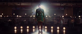Movie still from “Bad Times at the El Royale” (2018), directed by Drew Goddard – A woman in a green dress standing in front of lights; Wide shot, Over the shoulder angle