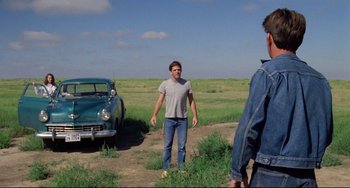 Movie still from “Badlands” (1973), directed by Terrence Malick – A man standing next to a car in a grassy field; Wide shot, Over the shoulder angle