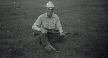 Movie still from “Badlands” (1973), directed by Terrence Malick – A man sitting in the grass holding a rifle; Medium shot, High angle