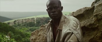 Movie still from “Blood Diamond” (2006), directed by Edward Zwick – A man in a tan shirt is posing for a picture on top of a mountain; Close Up shot, Low angle