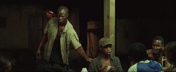 Movie still from “Blood Diamond” (2006), directed by Edward Zwick – A man standing next to a man sitting on a chair; Medium shot, Over the shoulder angle