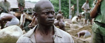 Movie still from “Blood Diamond” (2006), directed by Edward Zwick – A black man with a beard is standing in front of a group of people; Close Up shot, Low angle