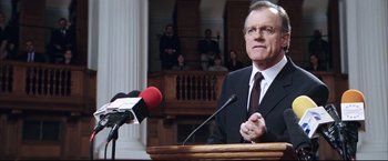 Movie still from “Blood Diamond” (2006), directed by Edward Zwick – A man in a suit and tie speaking at a podium; Medium shot, Low angle