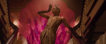 Movie still from “Booksmart” (2019), directed by Olivia Wilde – A man in a gold dress and black gloves standing in front of a pink background; Medium shot, Low angle