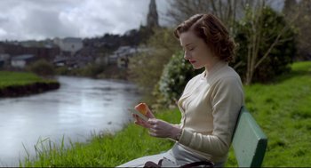 Movie still from “Brooklyn” (2015), directed by John Crowley – A woman sitting on a park bench looking at a piece of food; Medium shot, High angle