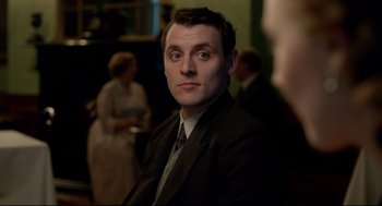 Movie still from “Brooklyn” (2015), directed by John Crowley – A man wearing a suit and tie sitting in front of a group of people; Close Up shot, Over the shoulder angle