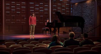 Movie still from “CODA” (2021), directed by Sian Heder – A woman standing in front of a grand piano in front of a group of people; Extreme Wide shot, High angle