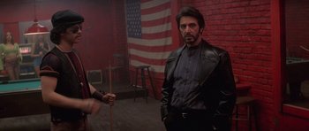 Movie still from “Carlito's Way” (1993), directed by Brian De Palma – A man in a leather jacket holding a stick in front of an american flag; Medium shot, Over the shoulder angle