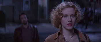 A woman with blonde curly hair wearing a brown jacket