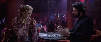 Movie still from “Carlito's Way” (1993), directed by Brian De Palma – Two people sitting at a table in front of a crowd; Medium shot, Over the shoulder angle