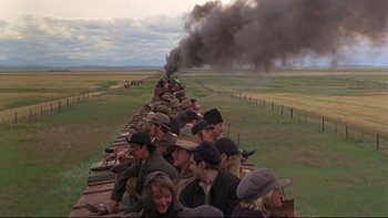 Movie still from “Days of Heaven” (1978), directed by Terrence Malick – A group of people riding on the back of a train on the tracks; Extreme Wide shot, High angle