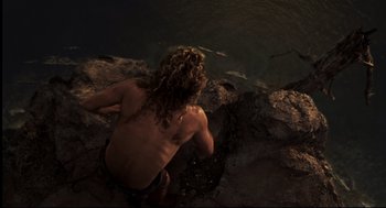 Movie still from “Cast Away” (2000), directed by Robert Zemeckis – A man with long curly hair sitting on a rock by a body of water; Wide shot, Overhead angle