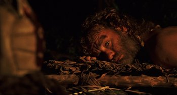 Movie still from “Cast Away” (2000), directed by Robert Zemeckis – A man with long hair and a beard laying on top of a pile of branches; Close Up shot, High angle