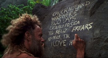 Movie still from “Cast Away” (2000), directed by Robert Zemeckis – A man writing on a rock with chalk on it; Wide shot, High angle