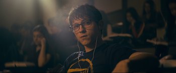 Movie still from “Cherry” (2021), directed by Joe Russo – A young man wearing glasses and a t - shirt with headphones; Close Up shot, Over the shoulder angle