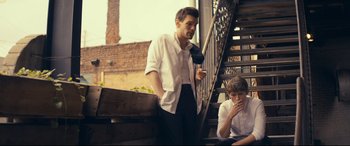 Movie still from “Cherry” (2021), directed by Joe Russo – A man standing next to a man sitting on steps; Medium shot, Low angle