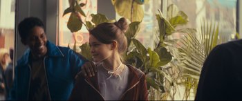 Movie still from “Cherry” (2021), directed by Joe Russo – A young girl smiles while standing in front of a plant; Medium shot, Over the shoulder angle