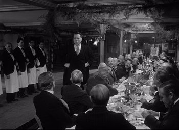 Movie still from “Citizen Kane” (1941), directed by Orson Welles – A man standing in front of a group of people at a dinner table; Medium shot, Over the shoulder angle