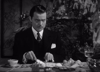 Movie still from “Citizen Kane” (1941), directed by Orson Welles – A black and white photo of a man in a suit cutting food; Medium shot, High angle