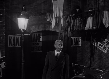 Movie still from “Citizen Kane” (1941), directed by Orson Welles – An older man in a suit standing in front of posters; Wide shot, High angle