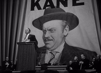 Movie still from “Citizen Kane” (1941), directed by Orson Welles – A black and white photo of a man speaking at a podium in front of a poster of john wayne; Medium shot, Low angle
