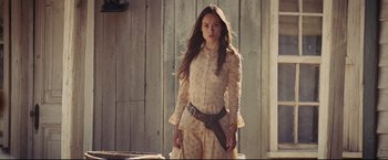 Movie still from “Cowboys & Aliens” (2011), directed by Jon Favreau – A beautiful young woman holding a gun while standing next to a building; Medium shot, Low angle
