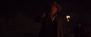 Movie still from “Cowboys & Aliens” (2011), directed by Jon Favreau – A man in a hat holding a knife in a dark room; Medium shot, Over the shoulder angle