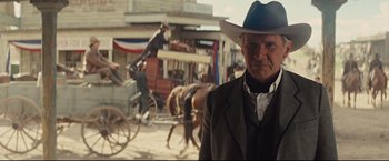 Movie still from “Cowboys & Aliens” (2011), directed by Jon Favreau – An older man in a suit and a cowboy hat stands in front of horse drawn carriages in an old western town; Medium shot, Over the shoulder angle