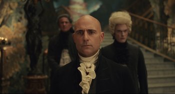 Movie still from “Cruella” (2021), directed by Craig Gillespie – A man with a bald head in front of two other men; Medium shot, Over the shoulder angle