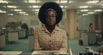 Movie still from “Cruella” (2021), directed by Craig Gillespie – A woman with a large afro sitting at a table; Medium shot, Over the shoulder angle