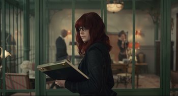 Movie still from “Cruella” (2021), directed by Craig Gillespie – A woman with red hair holding a book in her hands; Medium shot, Over the shoulder angle