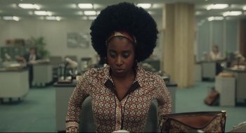 Movie still from “Cruella” (2021), directed by Craig Gillespie – A woman in a brown and white shirt is looking down at a book; Medium shot, Over the shoulder angle