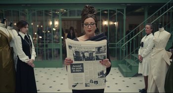 Movie still from “Cruella” (2021), directed by Craig Gillespie – A woman in a black top is holding a newspaper; Medium shot, High angle