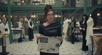 Movie still from “Cruella” (2021), directed by Craig Gillespie – A woman in a black dress holding a newspaper in a room; Medium shot, Over the shoulder angle