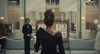 Movie still from “Cruella” (2021), directed by Craig Gillespie – A woman wearing a turban stands in front of a man in a suit; Medium shot, Over the shoulder angle