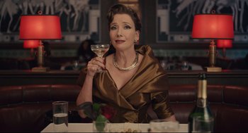 Movie still from “Cruella” (2021), directed by Craig Gillespie – A woman sitting at a table holding a wine glass; Medium shot, Over the shoulder angle