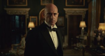 Movie still from “Cruella” (2021), directed by Craig Gillespie – A bald man in a black suit and a bow tie; Medium shot, Low angle
