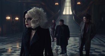 Movie still from “Cruella” (2021), directed by Craig Gillespie – A woman in a black coat standing in front of a staircase; Medium shot, Over the shoulder angle