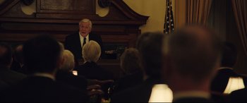 Movie still from “Dark Waters” (2019), directed by Todd Haynes – An older man in a black suit is sitting in front of an audience; Medium shot, Over the shoulder angle