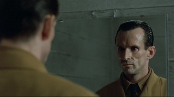 Movie still from “Downfall” (2004), directed by Oliver Hirschbiegel – A man looking at himself in the mirror in front of a wall; Close Up shot, Over the shoulder angle