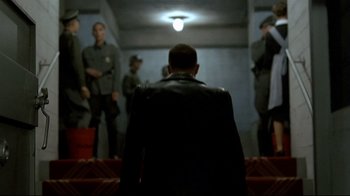 Movie still from “Downfall” (2004), directed by Oliver Hirschbiegel – A man wearing a leather jacket is sitting in front of a group of men; Wide shot, Over the shoulder angle