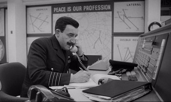 Movie still from “Dr. Strangelove or: How I Learned to Stop Worrying and Love the Bomb” (1964), directed by Stanley Kubrick – A man sitting at a desk on a phone; Medium shot, High angle