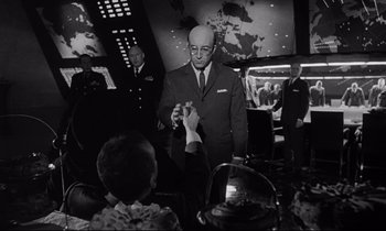 Movie still from “Dr. Strangelove or: How I Learned to Stop Worrying and Love the Bomb” (1964), directed by Stanley Kubrick – A man in a suit and tie holding a cell phone in front of other men; Medium shot, Low angle