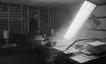Movie still from “Dr. Strangelove or: How I Learned to Stop Worrying and Love the Bomb” (1964), directed by Stanley Kubrick – A black and white photo of a man standing in front of a table; Wide shot, High angle