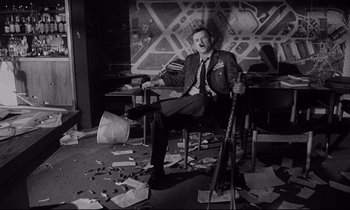 Movie still from “Dr. Strangelove or: How I Learned to Stop Worrying and Love the Bomb” (1964), directed by Stanley Kubrick – A man sitting in a chair in a room with a lot of papers on the floor; Wide shot, Low angle