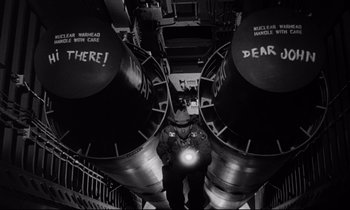 Movie still from “Dr. Strangelove or: How I Learned to Stop Worrying and Love the Bomb” (1964), directed by Stanley Kubrick – A man standing next to two large pipes in a room; Medium shot, Overhead angle