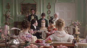 Movie still from “Emma.” (2020), directed by Autumn de Wilde – A group of people standing around a table with a cake on it; Medium shot, Over the shoulder angle
