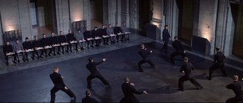 Movie still from “Equilibrium” (2002), directed by Kurt Wimmer – A group of men in black suits are standing in front of a row of chairs; Wide shot, High angle
