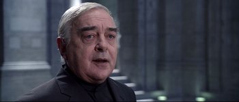 Movie still from “Equilibrium” (2002), directed by Kurt Wimmer – An older man in a black suit is standing in front of a staircase; Close Up shot, Over the shoulder angle