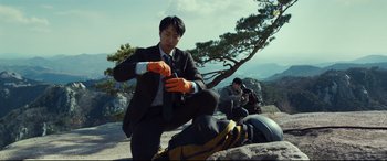 Movie still from “Decision to Leave” (2022), directed by Park Chan-wook – A man in a black suit and orange safety gloves; Medium shot, Low angle